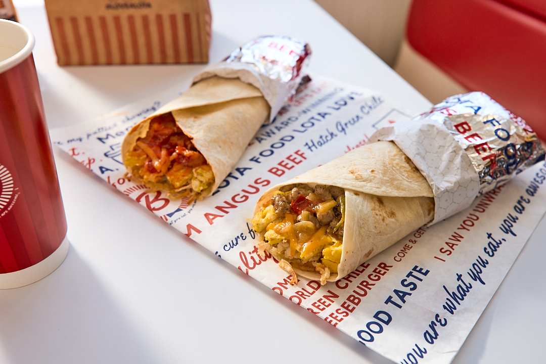 Two Blake's breakfast burritos in wrappers on top of a table with cup and bag
