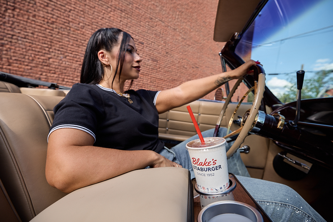 Low rider 15754 Woman with dark hair in black shirt sitting in a lowrider with a Blake's drink cup in the cupholder