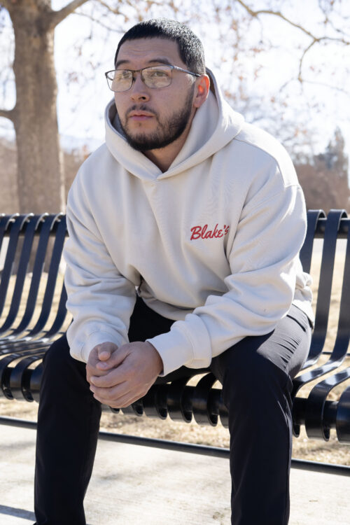 Man sitting on park bench wearing an off-white hoodie with the word Blake's embroidered on it in red.