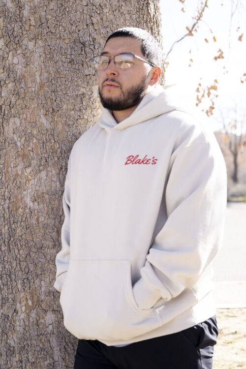 Man leaning against tree wearing an off-white hoodie with the word Blake's embroidered on it in red.